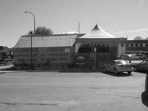 The Klondike Airways building appears as a wood frame building in a 1906 photo. It is located on a site previously occupied by a tent frame bakery. The building was later covered in galvanized metal and used as a warehouse.